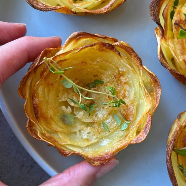 A hand holds a crispy, golden-brown potato rose, garnished with fresh thyme and displayed on a light blue plate with more elegant potato roses in the background.