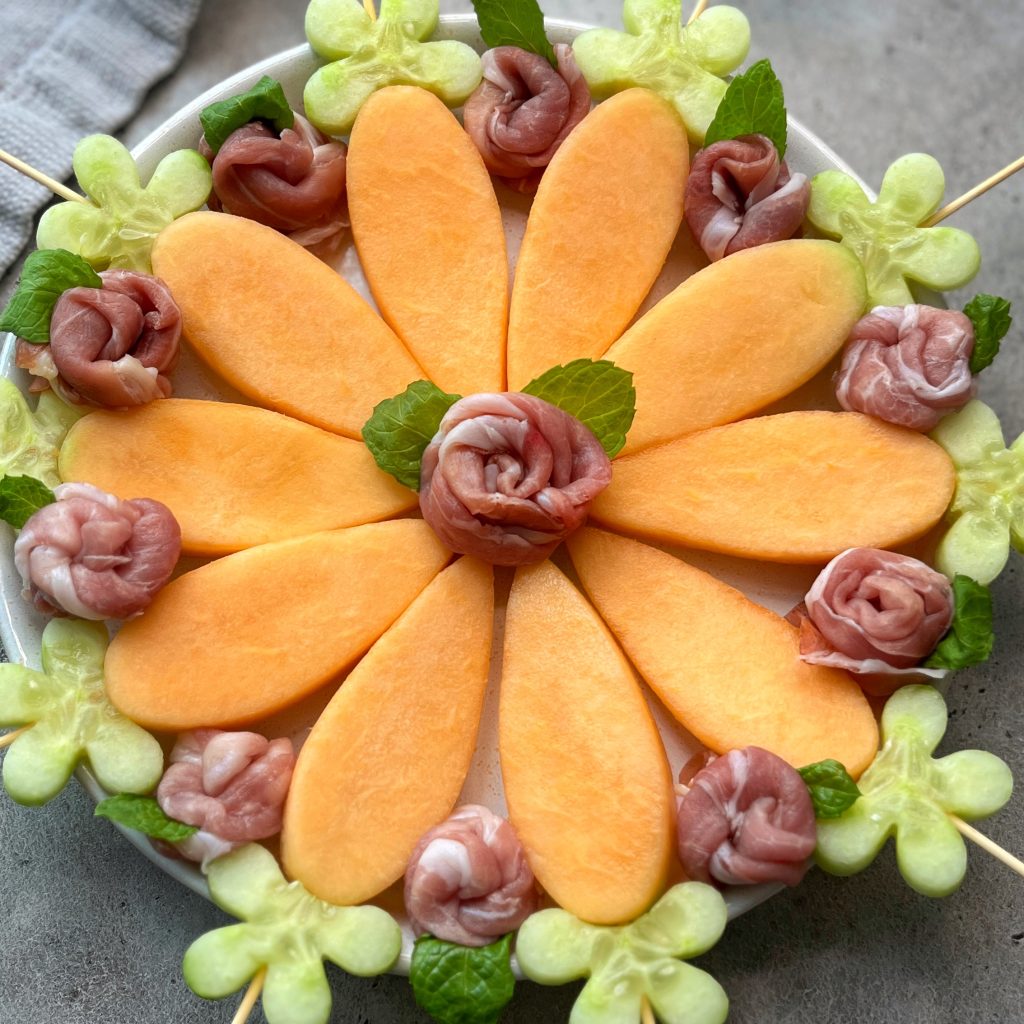 A platter arranged with prosciutto and melon: cantaloupe slices as flower petals, prosciutto roses in the center and between petals, green melon flowers on the edge, and mint leaves for garnish.