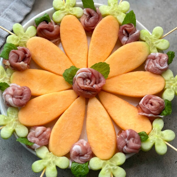 A platter arranged with prosciutto and melon: cantaloupe slices as flower petals, prosciutto roses in the center and between petals, green melon flowers on the edge, and mint leaves for garnish.
