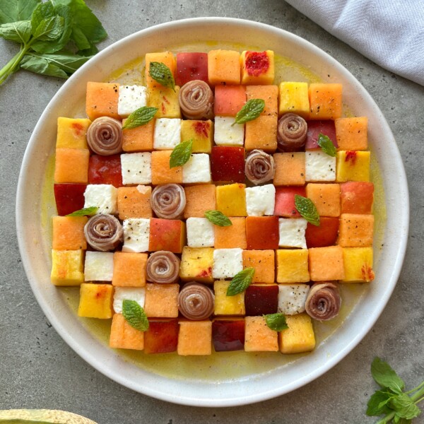 A plate with a colorful prosciutto melon salad featuring neatly cubed melon, watermelon, mozzarella, and rolled prosciutto, garnished with fresh mint leaves and ground pepper, all arranged on a light sauce.