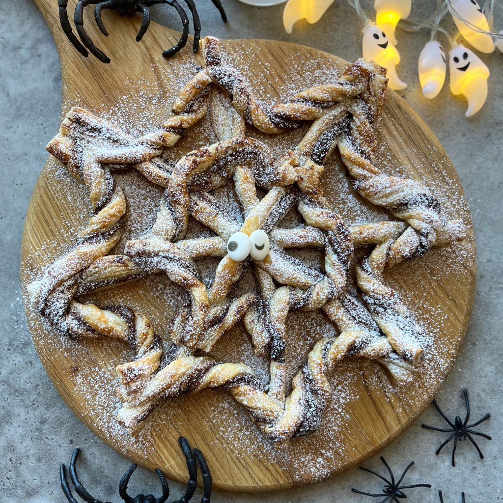 A spiderweb-shaped pastry dusted with powdered sugar sits on a round wooden board, decorated with candy eyes, surrounded by plastic spiders and glowing ghost lights—a festive Halloween treat perfect for creative food recipes and fun party bites.