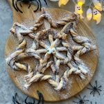 A spiderweb-shaped pastry dusted with powdered sugar sits on a round wooden board, decorated with candy eyes, surrounded by plastic spiders and glowing ghost lights—a festive Halloween treat perfect for creative food recipes and fun party bites.