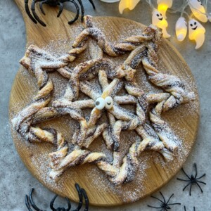 A spiderweb-shaped pastry dusted with powdered sugar sits on a round wooden board, decorated with candy eyes, surrounded by plastic spiders and glowing ghost lights—a festive Halloween treat perfect for creative food recipes and fun party bites.