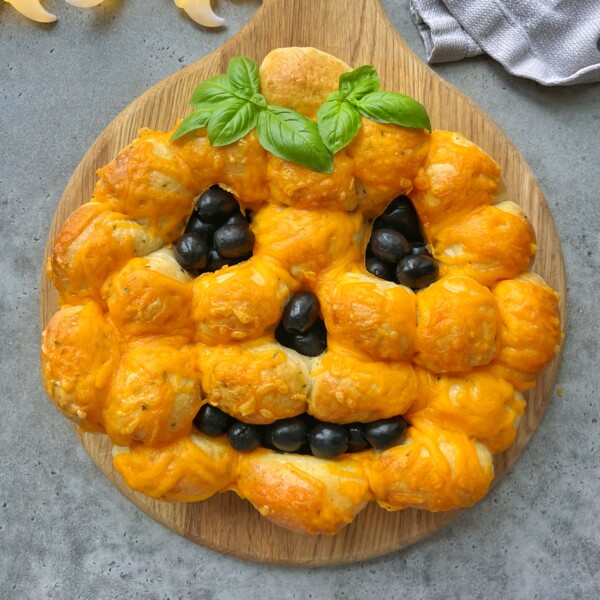 Pumpkin-shaped pull-apart bread, topped with melted cheddar cheese and decorated with black olive bites for the face and fresh basil as a stem, is a fun dish to add to your collection of festive food recipes. Served on a round wooden board.