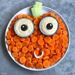 A round plate arranged with carrot slices, two cups of hummus topped with olives for eyes, cucumber pieces for a stem, and a curved vegetable smile creates a pumpkin face—perfect for fun, healthy bites and festive food recipes.