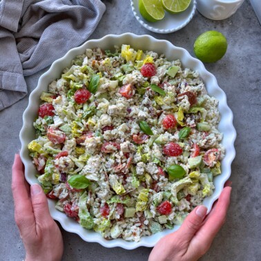 A person holds a round dish filled with a colorful rice chicken salad made of lettuce, cherry tomatoes, avocado, and fresh basil. Lime halves and a kitchen towel are nearby on a gray surface.