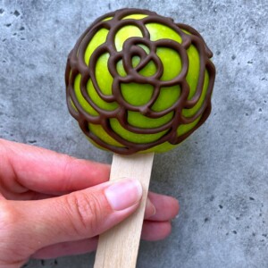 A hand holds a green apple on a wooden stick, decorated with an intricate swirl pattern of drizzled dark chocolate—an irresistible treat that brilliantly combines health and indulgence in every bite, against a gray textured background.