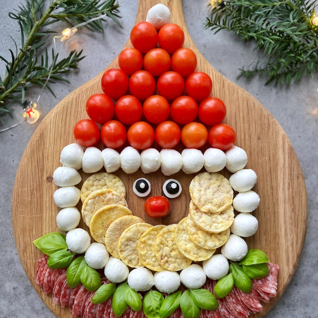 A festive Santa Charcuterie Board features Santa’s face made from cherry tomatoes, mozzarella balls, rice crackers, basil, salami, and olives on a wooden board, surrounded by pine branches and twinkling string lights.