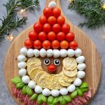 A festive Santa Charcuterie Board features Santa’s face made from cherry tomatoes, mozzarella balls, rice crackers, basil, salami, and olives on a wooden board, surrounded by pine branches and twinkling string lights.