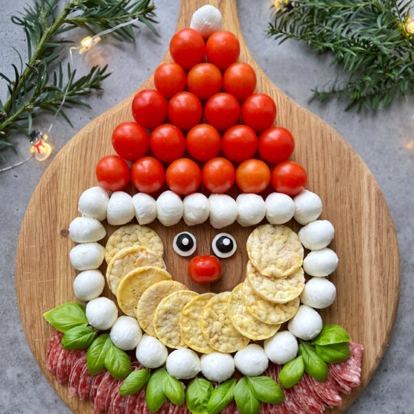 A festive Santa Charcuterie Board features Santa’s face made from cherry tomatoes, mozzarella balls, rice crackers, basil, salami, and olives on a wooden board, surrounded by pine branches and twinkling string lights.