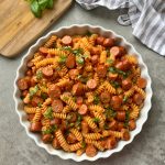 A white round dish filled with Sausage Pasta—rotini, sliced sausages, and tomato sauce—garnished with fresh basil. A wooden board with basil and a striped cloth rest nearby on a gray countertop.