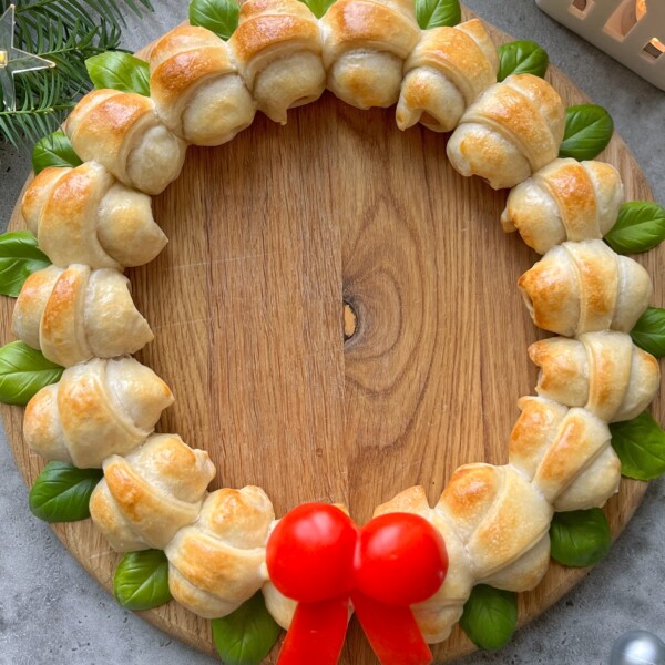 A festive Sausage Roll Wreath made of small golden croissants, fresh green basil leaves, and two red cherry tomatoes with a pepper slice bow, arranged on a round wooden board.