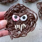 A hand holds a round chocolate treat with tangled swirls, two candy eyes—one bloodshot—and white candy sprinkles arranged as fangs, resembling a monster face.