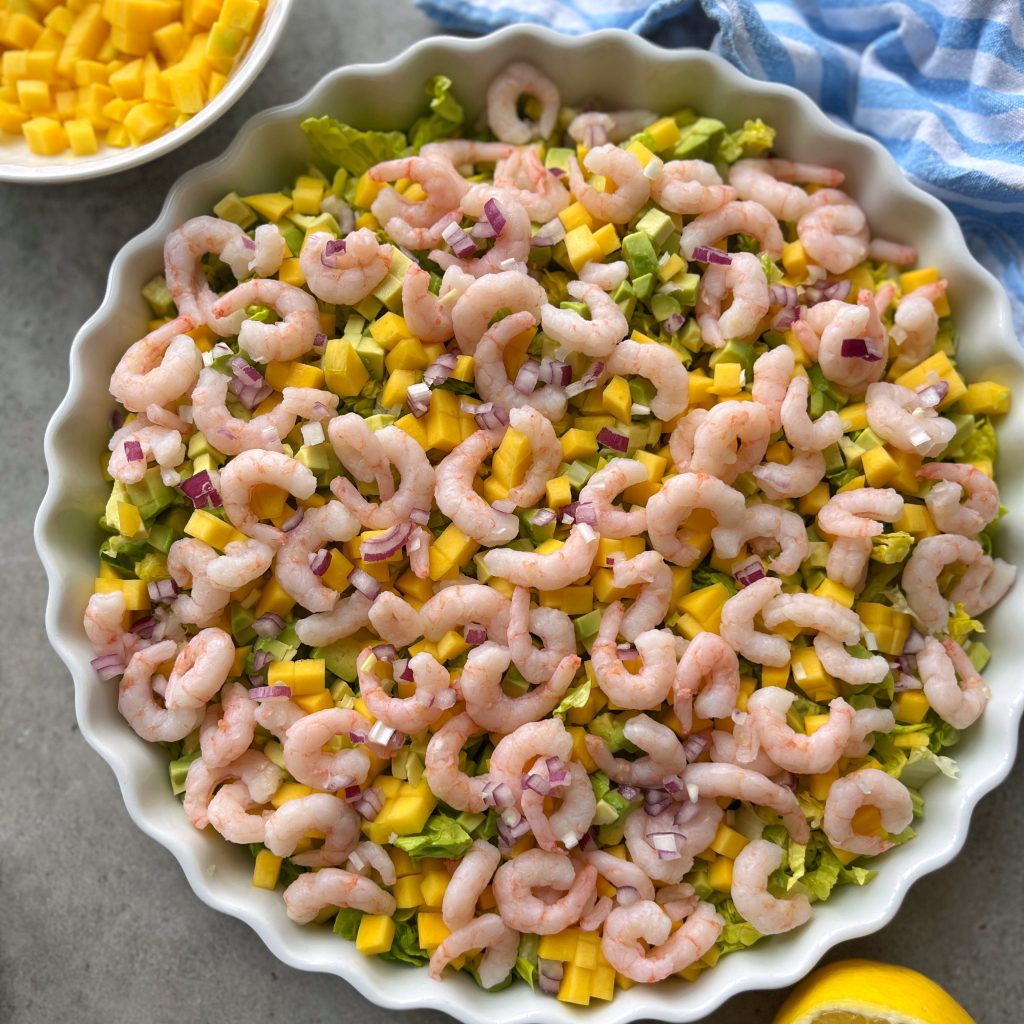 A round dish filled with a vibrant shrimp cocktail salad featuring diced mango, avocado, chopped red onion, and lettuce. Nearby are a bowl of mango cubes and a lemon wedge on a gray surface with a blue striped cloth.