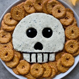 A spooky food dish featuring a skull-shaped dip with black olive eyes and mouth, surrounded by round crackers arranged in a circle—perfect for creative bites and festive recipes.