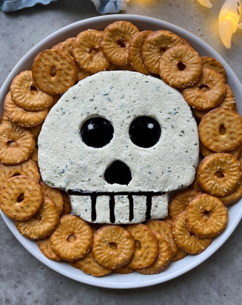 A spooky food dish featuring a skull-shaped dip with black olive eyes and mouth, surrounded by round crackers arranged in a circle—perfect for creative bites and festive recipes.