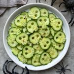 Cucumber slices carved to look like skulls are arranged on a white plate, sprinkled with red pepper flakes for a healthy twist. Plastic black spiders surround these festive bites, making them perfect for Halloween-themed food recipes.