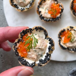A hand holds a flower-shaped pastry cup topped with creamy filling, orange fish roe, black sesame seeds, and a sprig of dill—delicious bites perfect for health-conscious food recipes. More similar appetizers rest on a round white platter behind.