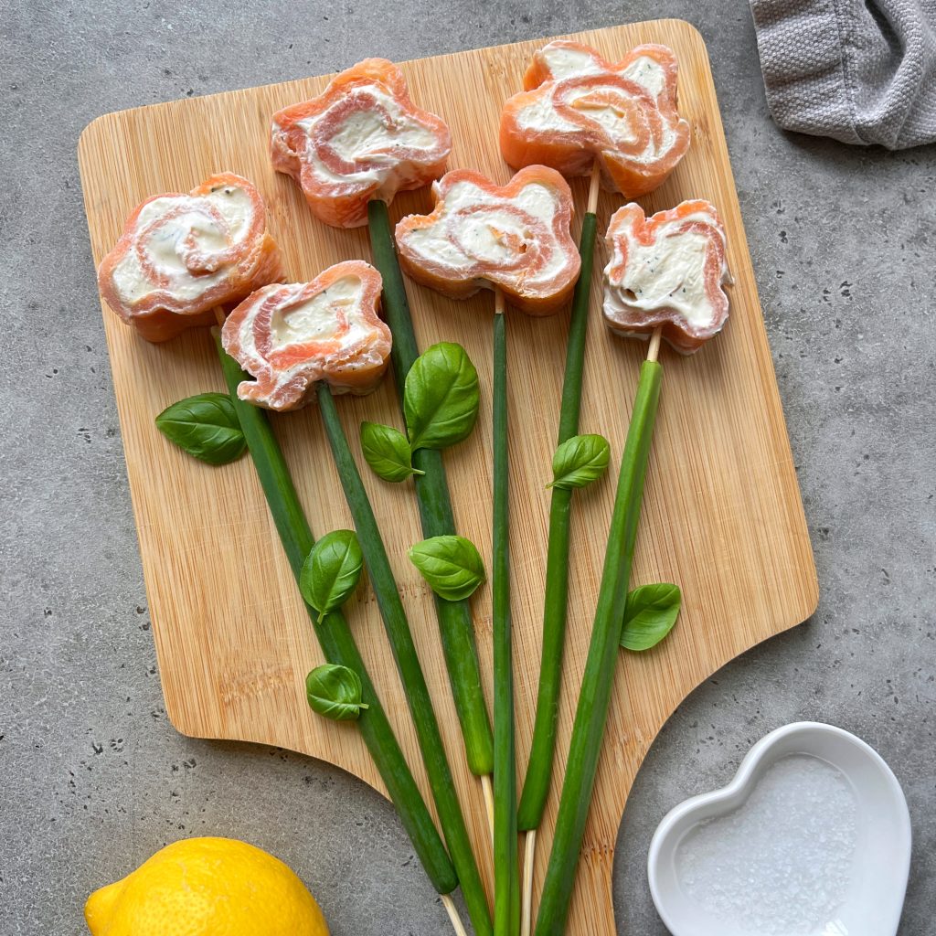 Sushi rolls shaped like flowers with cream cheese centers are arranged on a wooden board, resembling a smoked salmon roulade bouquet. Green onions and basil leaves form stems and leaves. A lemon and a heart-shaped bowl are nearby.