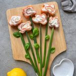 Sushi rolls shaped like flowers with cream cheese centers are arranged on a wooden board, resembling a smoked salmon roulade bouquet. Green onions and basil leaves form stems and leaves. A lemon and a heart-shaped bowl are nearby.