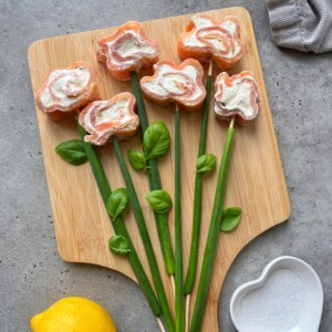 Sushi rolls shaped like flowers with cream cheese centers are arranged on a wooden board, resembling a smoked salmon roulade bouquet. Green onions and basil leaves form stems and leaves. A lemon and a heart-shaped bowl are nearby.