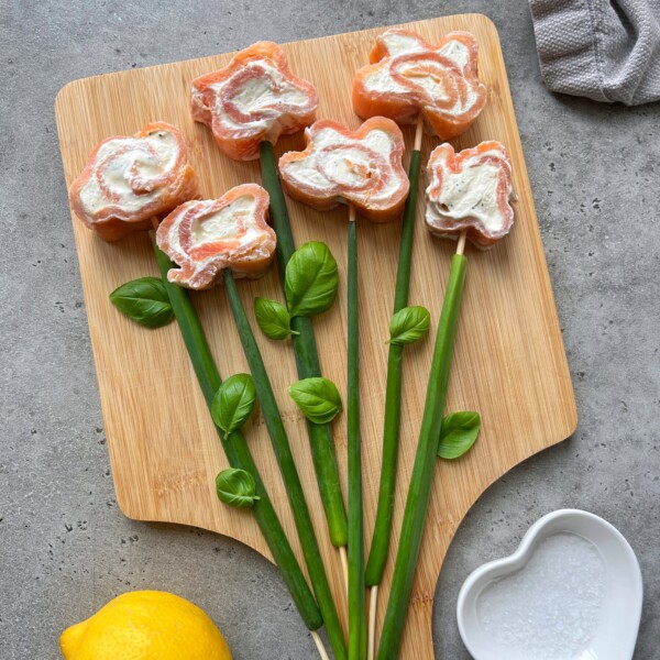 Sushi rolls shaped like flowers with cream cheese centers are arranged on a wooden board, resembling a smoked salmon roulade bouquet. Green onions and basil leaves form stems and leaves. A lemon and a heart-shaped bowl are nearby.