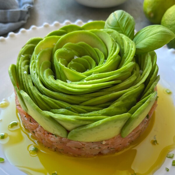 A neatly arranged avocado rose sits atop a base of finely chopped smoked salmon tartare, garnished with basil leaves and drizzled with olive oil on a white plate.