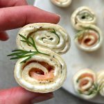 A hand holds a pinwheel-shaped appetizer—tortilla rolled with cream cheese, smoked salmon, herbs, and topped with dill. These healthy bites are perfect for parties and make an elegant addition to your food recipes or appetizer dishes.