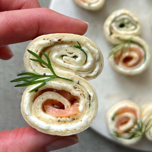 A hand holds a pinwheel-shaped appetizer—tortilla rolled with cream cheese, smoked salmon, herbs, and topped with dill. These healthy bites are perfect for parties and make an elegant addition to your food recipes or appetizer dishes.