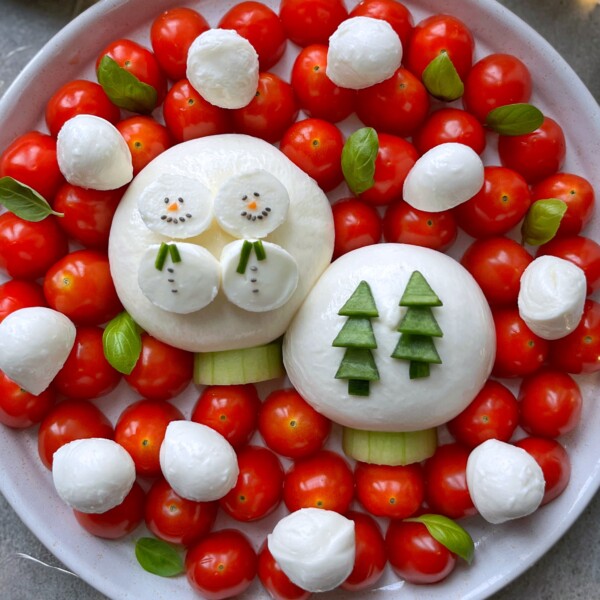 A Snow Globe Burrata Platter arranged with cherry tomatoes and mozzarella balls, featuring two large mozzarella rounds decorated as a snowman and two trees with cucumber details, surrounded by basil leaves.