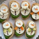 Slices of bread topped with cheese, cucumbers, and vegetables arranged to look like snowmen with carrot noses, olive eyes, and scarf decorations—these festive bites make fun and healthy food recipes for any celebration.