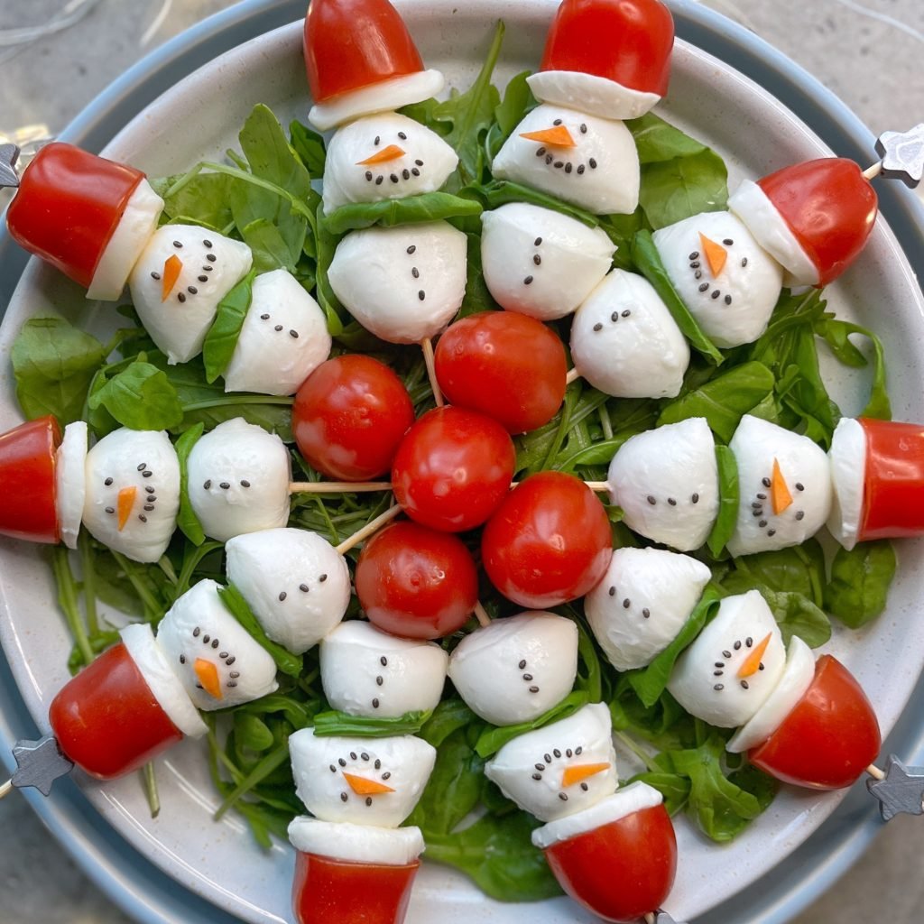 A plate of salad arranged with mozzarella and tomato snowmen, each with black dots for eyes and mouth, and a carrot nose, on a bed of greens—fun bites that make healthy food recipes festive. Cherry tomatoes form Santa hats at the center of the dish.