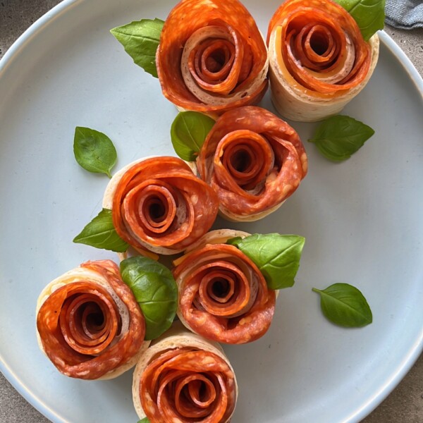 Six rolled slices of pepperoni arranged as roses on a plate with small green basil leaves, resembling a bouquet of flowers—perfect for creative food recipes or appetizer bites.