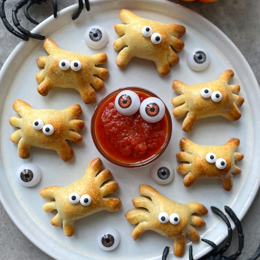 A round white plate with eight Spider Sausage Rolls, each shaped like a crab and topped with candy eyes, arranged around a bowl of red sauce with two candy eyes. Fake eyeballs and decorative black claws accent the plate.