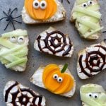 Slices of bread with cream cheese are decorated for Halloween as mummies, spiders, and pumpkins using cucumber, yellow vegetables, balsamic glaze, and candy eyes. Plastic spiders are placed around the snacks.