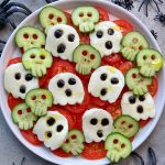 A plate of sliced tomatoes, cucumbers shaped like skulls, and mozzarella ghosts with black olive eyes makes a spooky, healthy Halloween dish. Plastic spiders are scattered around for extra fun—a perfect addition to festive food recipes!.