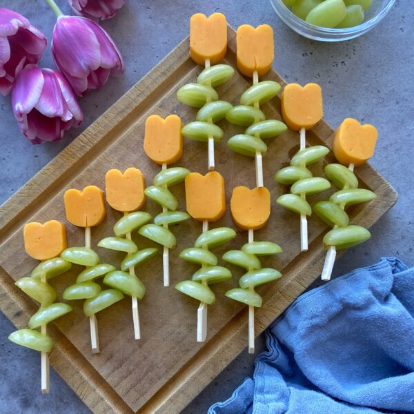 Tulip-shaped cheddar cheese slices top green grape skewers, creating colorful, healthy bites on a wooden cutting board. Pink tulips and a blue napkin complete this vibrant scene—perfect for inspiring your next food recipes.