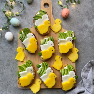 Decorated toasts topped with white and yellow frosting shaped like chicks and green avocado slices, arranged on a wooden board with yellow chick-shaped marshmallows. These charming bites make fun, festive dishes for springtime food recipes. Fairy lights glow in the background.