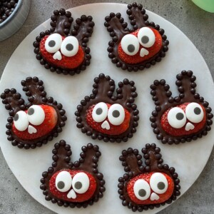 Seven Strawberry Bunnies made with halved strawberries, chocolate ears and borders, large candy eyes, and tiny white candy noses are arranged on a round white plate.