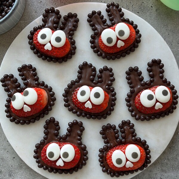 Seven Strawberry Bunnies made with halved strawberries, chocolate ears and borders, large candy eyes, and tiny white candy noses are arranged on a round white plate.