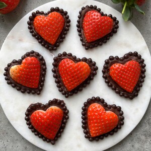 Seven heart-shaped strawberry halves are placed on a round white marble plate, each outlined with a border of chocolate dots. This delightful arrangement combines elegance and health for a romantic dessert perfect for food recipes and special bites.