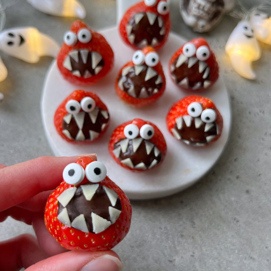 A hand holds a strawberry decorated like a monster with candy eyes and white chocolate teeth. More monster bites are arranged on a plate in the background, surrounded by ghost-shaped lights, adding fun to your food recipes.