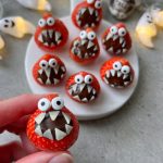 A hand holds a strawberry decorated to look like a monster with chocolate filling, candy eyes, and white chocolate teeth. These playful bites make fun food for parties, with more monster strawberries on a plate surrounded by ghost decorations.