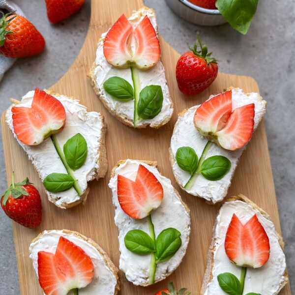 Strawberry crostini featuring bread slices topped with cream cheese, strawberry halves shaped like tulips, and basil leaves as stems are artfully arranged on a wooden board with fresh strawberries nearby.