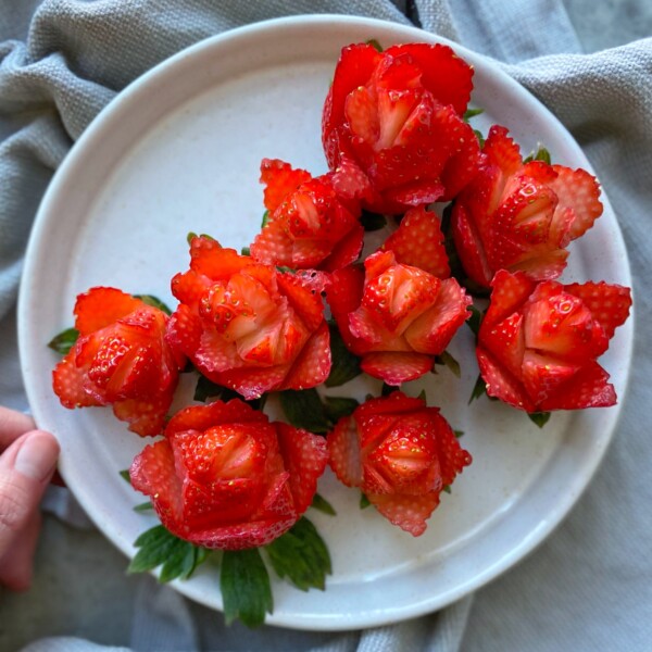 A white plate holds strawberries carved to resemble blooming red roses, with green strawberry leaves beneath them—an inspiring idea for creative food recipes. The plate rests on a gray cloth, and a hand is partially visible on the left edge.