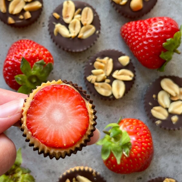 A hand holds a chocolate cup topped with a sliced strawberry, reminiscent of a strawberry Snickers treat. Other chocolate cups with peanuts and whole strawberries are arranged on a gray surface in the background.