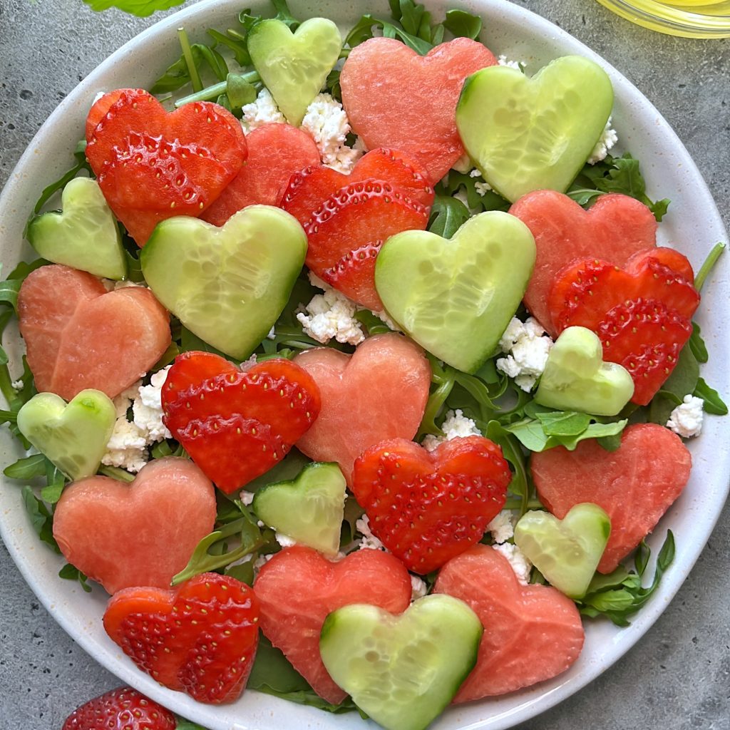 A delightful strawberry watermelon salad featuring heart-shaped pieces of watermelon, cucumber, and strawberries on a bed of greens, with crumbled white cheese scattered throughout.