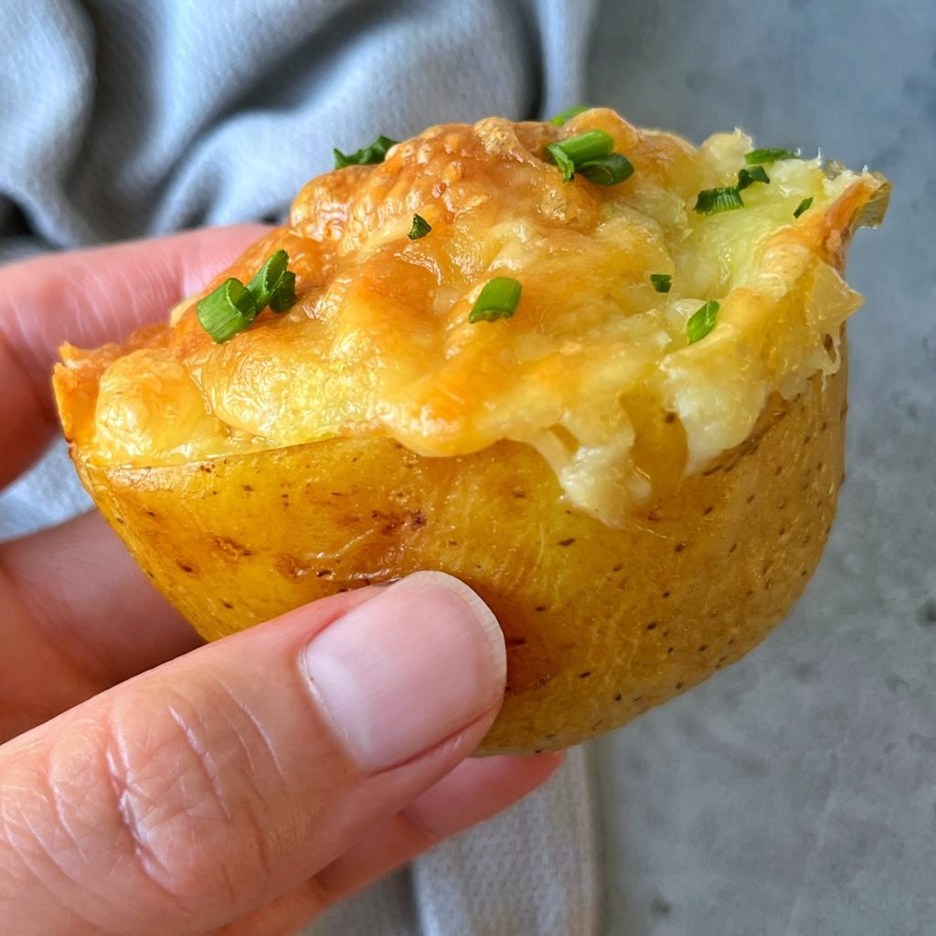 A hand holding a baked potato half topped with melted cheese, tuna, and chopped chives, against a blurred gray background.