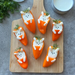 Stuffed Mini Peppers are transformed into adorable bunny faces—orange bell pepper halves filled with creamy white dip, then decorated with dill, black sesame seeds, and marshmallows on a wooden cutting board.