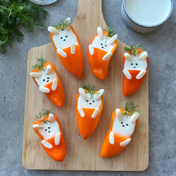 Stuffed Mini Peppers are transformed into adorable bunny faces—orange bell pepper halves filled with creamy white dip, then decorated with dill, black sesame seeds, and marshmallows on a wooden cutting board.